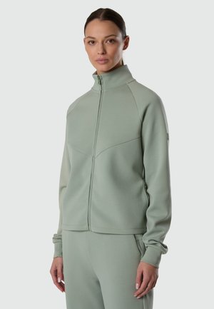 Frau trägt eine salbeigrüne Jacke mit Reißverschluss und hohem Kragen sowie passende Hose, steht vor einem hellgrauen Hintergrund.