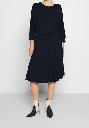 Kobieta w czarnej sukience z długim rękawem, z asymetrycznym dołem oraz białych botkach za kostkę z czarnymi ostrymi czubkami i obcasami.