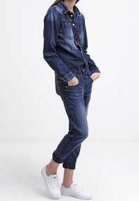 Persona vestida con una chaqueta de mezclilla azul oscura y jeans con los puños remangados, de pie con las manos en los bolsillos y zapatillas blancas.