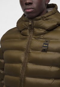 Gros plan d'une personne portant une doudoune vert olive avec une fermeture éclair frontale et un petit patch de marque sur la poitrine.