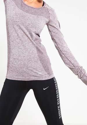 Femme en t-shirt violet à manches longues et leggings noirs Nike, s'étirant avec les bras tendus devant un fond blanc uni.