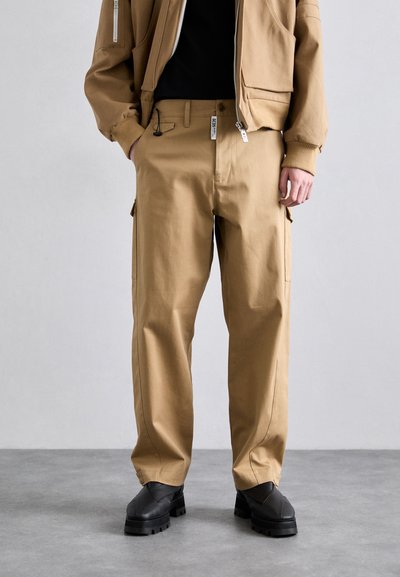 Pantaloni cargo bej cu o croială lejeră, având buzunare laterale, o textură netedă și un material durabil. Asortați cu pantofi negri, masivi.