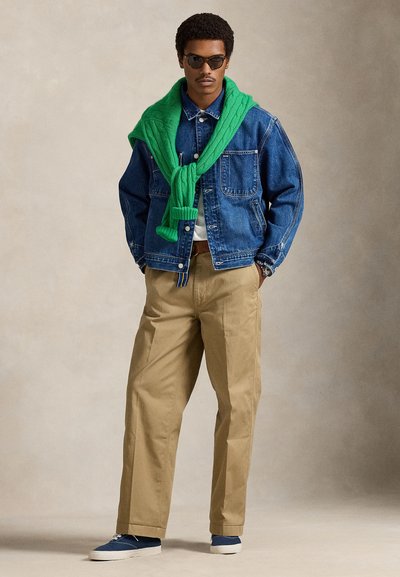 Τζιν μπουφάν με μπροστινές τσέπες, πράσινο πλεκτό πουλόβερ περασμένο πάνω από τους ώμους, μπεζ παντελόνια και μπλε αθλητικά παπούτσια. Το μοντέλο φορά γυαλιά ηλίου.