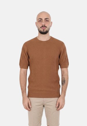 Uomo con barba e tatuaggio che indossa un maglione marrone a maniche corte con texture e pantaloncini beige, in piedi davanti a uno sfondo chiaro e semplice.