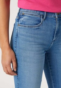 Vaqueros de denim azul claro con un corte slim, que cuentan con remaches de metal en tono dorado y costuras en contraste, destacando los bolsillos y una textura suave.