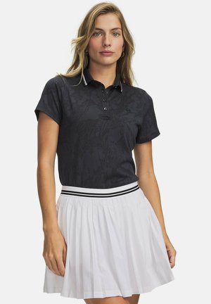 Vrouw met lang blond haar, gekleed in een zwart poloshirt met korte mouwen en een witte geplooide rok met gestreepte tailleband, staand tegen een witte achtergrond.