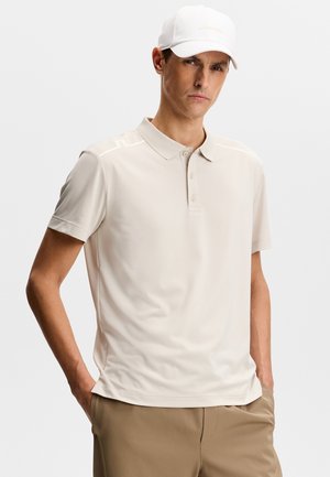 Mann mit heller Haut, der eine weiße Baseballkappe, ein beiges Poloshirt und braune Hosen trägt, steht mit den Händen in den Taschen vor weißem Hintergrund.