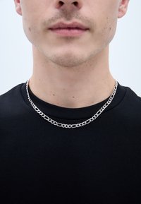 Suds sudraba metāla ķēde ar divām kārtām, kas aprīkota ar savstarpēji saistītām saitēm, kas valkāta virs melna krūšu apkakles krekla.