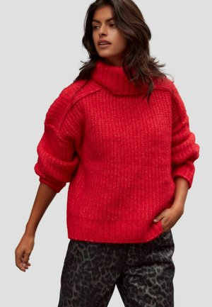 Fleckiger roter Pullover mit hohem, gefaltetem Kragen. Verfügt über ein geripptes Muster und übergroße Ärmel, kombiniert mit dunklen, gemusterten Hosen.