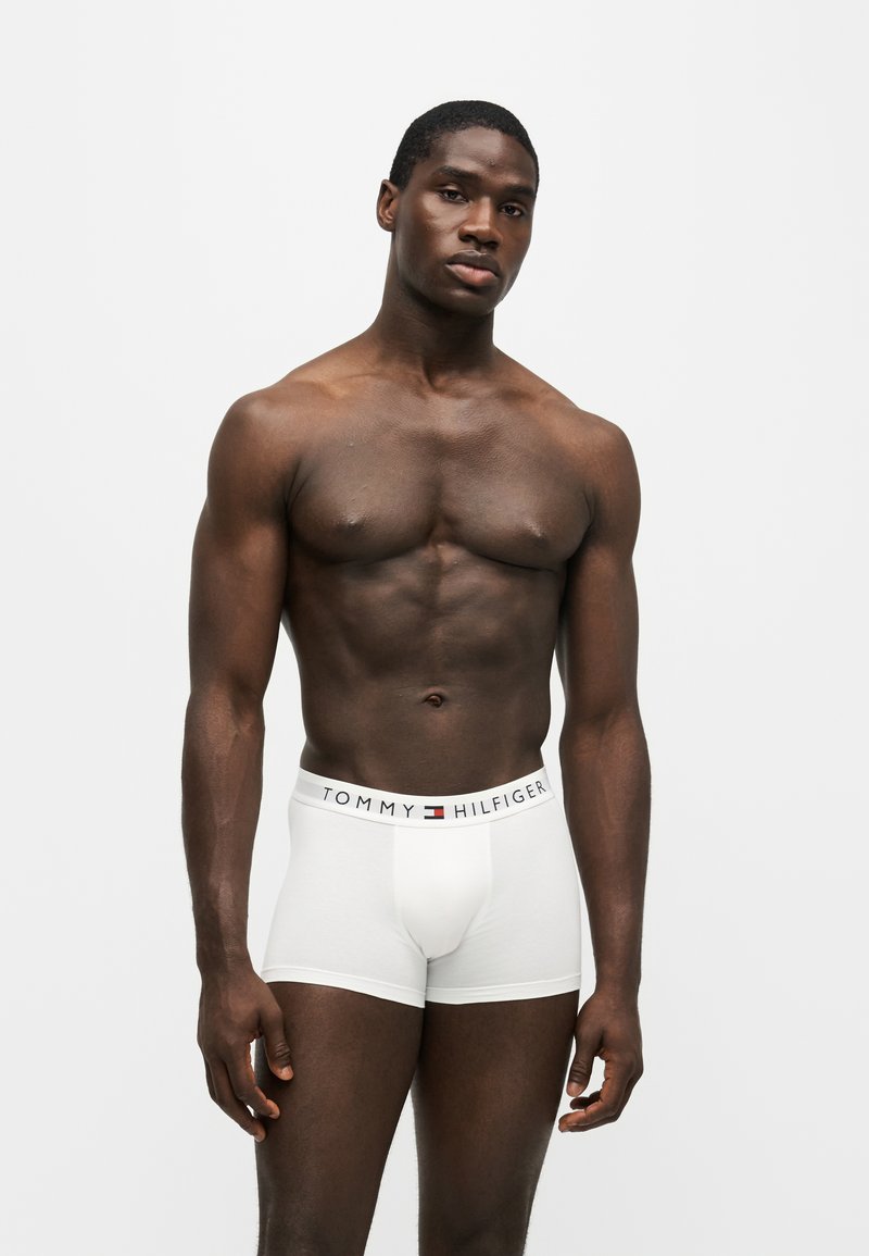 Muskelbepackter Mann steht, trägt weiße Tommy Hilfiger Boxershorts, vor einem schlichten hellgrauen Hintergrund mit neutralem Gesichtsausdruck.