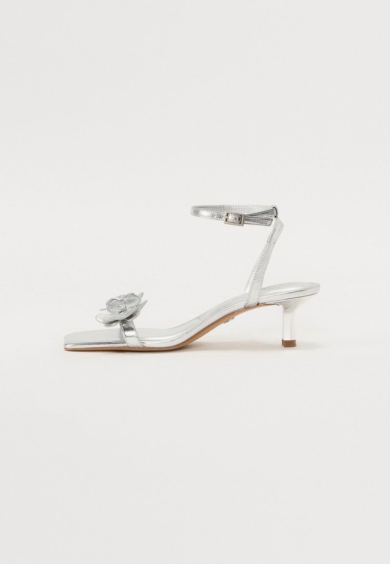 Hõbe metallik sandaalid madala kontsaga, säärepaela ja lillemotiiviga varvastepaelal; sile tekstuur ja elegantne disain täiustavad nende välimust.