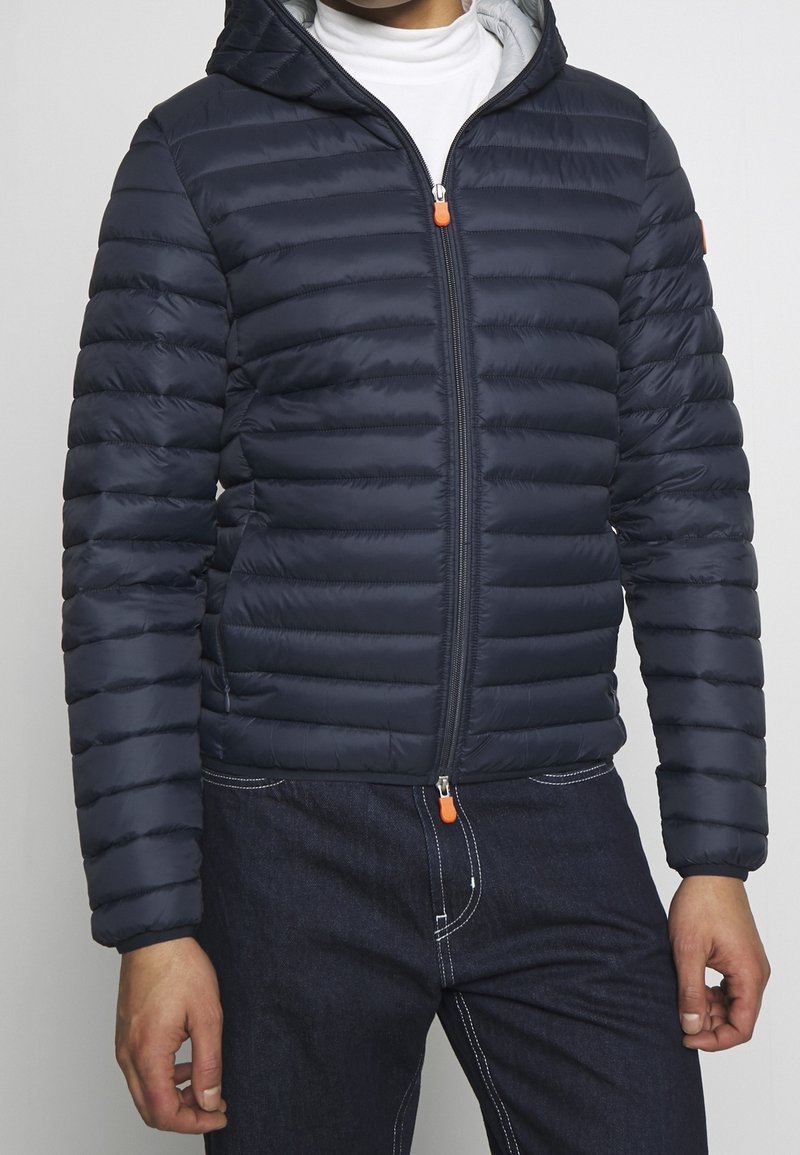Giubbotto a vento navy con sezioni trapuntate orizzontali, cappuccio e una linguetta arancione; realizzato in materiale leggero e resistente all'acqua.