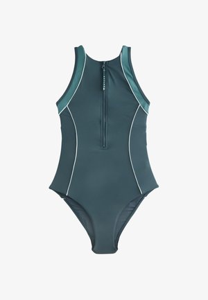 Athletische Frau trägt einen blauen Badeanzug mit Reißverschluss vorne und gelb getönte Sportsonnenbrille, steht mit den Händen in den Hüften vor klarem Himmel.