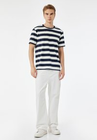 T-paita, jossa on tummansinisiä ja valkoisia raitoja, pyöreä kaula-aukko ja lyhyet hihat, yhdistettynä vaaleisiin suoriin housuihin ja valkoisiin lenkkareihin.