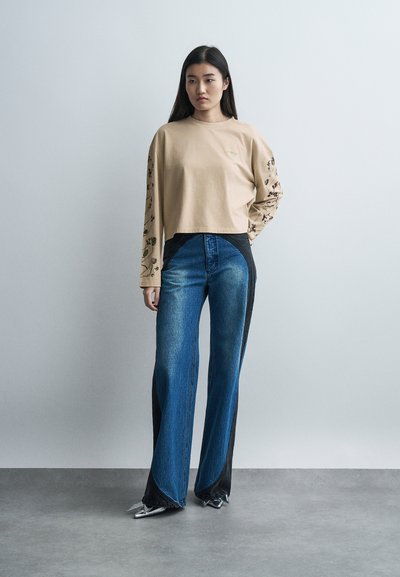 Νεαρή γυναίκα που στέκεται, φοράει μπλούζα με μακριά μανίκια σε μπεζ χρώμα με φλοράλ μοτίβα στα μανίκια, διχρωμία σε παντελόνι με φαρδιές μπατζάκια και ασημένια μυτερά παπούτσια.