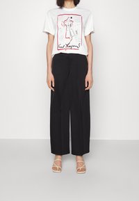 Bílý grafický tričko s designem s červenými akcenty, spárované s černými širokými kalhotami a světle hnědými sandály na páse.