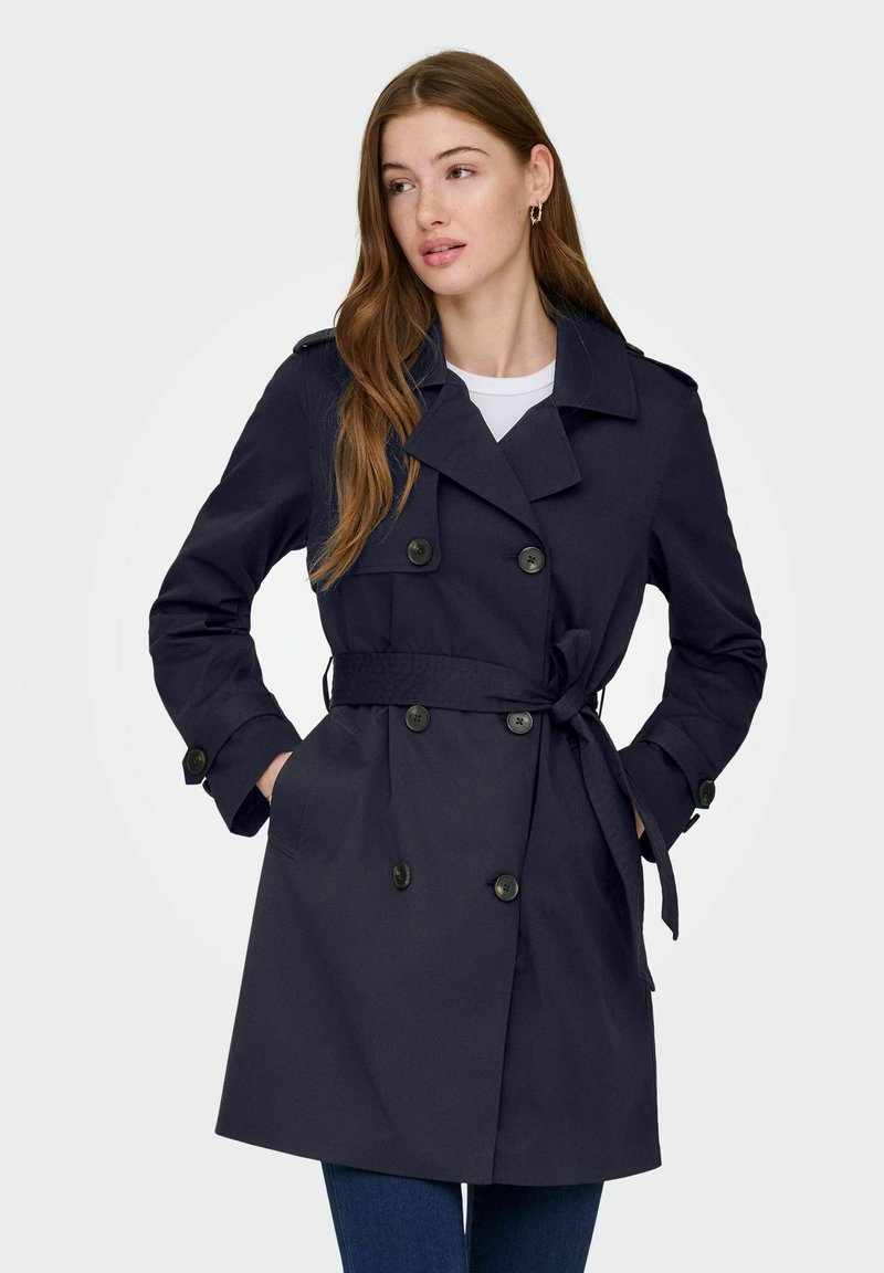 Femme aux longs cheveux bruns portant un trench-coat bleu marine croisé avec ceinture nouée à la taille et les mains dans les poches.