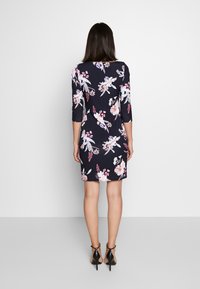 Nainen seisoo selkä cameraan päin, yllä tummansininen mekko, jossa on vaaleanpunaisia ja valkoisia kukkakuoseja, sekä mustat korkokengät yksinkertaista taustaa vasten.