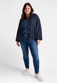 Granatowa lekka kurtka zapinana na zamek, noszona z dżinsową bluzką i niebieskimi rurkami z ozdobami, w połączeniu z białymi sneakerami.