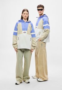 Színblokkokkal díszített széldzsekik bézs, világoskék és fehér színekben, félcipzárral, húzózsinóros kapucnival és elülső zsebekkel. Bő, széles szárú nadrágok, laza fazonban.