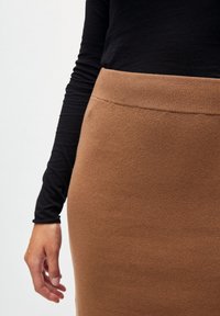 Persoon in een zwarte lange mouwtop en een high-waisted bruine gebreide rok, met een ontspannen hand aan de zijkant tegen een effen achtergrond.