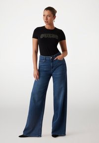 Tricou negru, ajustat, cu "GUESS" scris în cristale, asortat cu blugi albaștri cu talie înaltă și iarbă largă. Material texturat și finisare impecabilă.