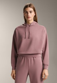 Jonge vrouw met lang, stijl haar, gekleed in een mauve capuchontrui en bijpassende joggingbroek, staand tegen een effen achtergrond.