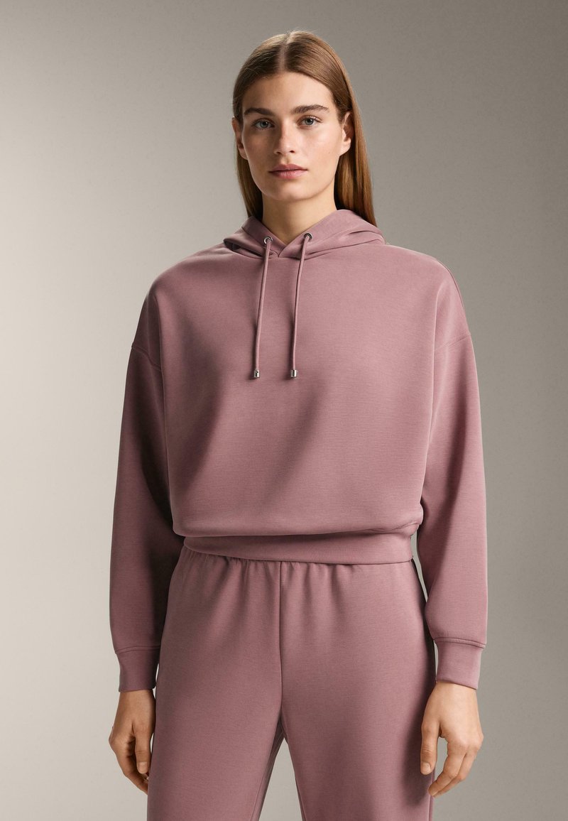 Jonge vrouw met lang, stijl haar, gekleed in een mauve capuchontrui en bijpassende joggingbroek, staand tegen een effen achtergrond.