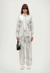 Silberne metallische Kleidung mit einem Hemd mit Knöpfen und lockeren Hosen, die eine glänzende Textur aufweisen. Eine schwarze Handtasche und Sonnenbrillen runden den Look ab.