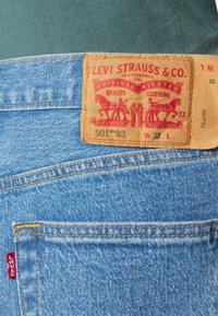 Blue denim jeans featuring a brown leather patch with red printing, back pocket detail, and yellow stitching. Classic fit with a faded look.