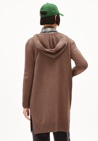 Brauner, langer Cardigan mit Kapuze, aus weichem Material. Verfügt über gerippte Bündchen und seitliche Schlitze. Wird über einem karierten Hemd getragen. Grüne Mütze sichtbar.