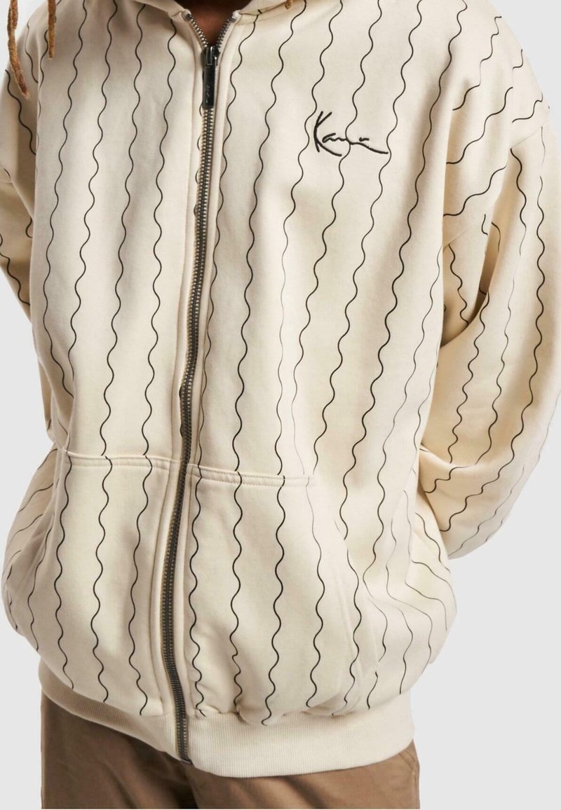 Beige zip-up jacka med vertikala vågiga svarta linjer och liten svart textlogotyp på vänster bröst, bärs över bruna byxor.