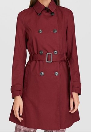 Kobieta nosząca bordowy, dwurzędowy trencz do kolan z paskiem w talii i zapinanymi na guziki mankietami.