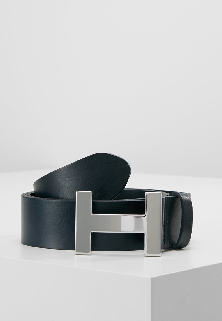 Ceinture en cuir noir roulée avec une grande boucle métallique argentée en forme de la lettre H sur une surface blanche.
