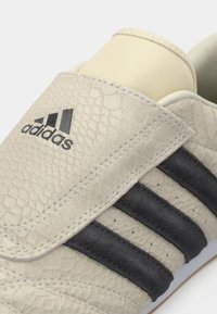 Μπεζ αθλητικό παπούτσι με υφή στην επάνω επιφάνεια, μαύρες σουέντ ρίγες και ανάγλυφο σχέδιο. Διαθέτει το λογότυπο της Adidas στο πλευρό και επενδεδυμένη γλώσσα.