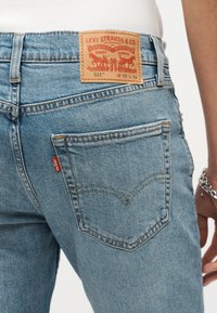 Φωτεινά γαλάζια τζιν Levi's 511 που φοριούνται από ένα άτομο, δείχνοντας την πίσω τσέπη, την κόκκινη ετικέτα της μάρκας και το δερμάτινο σημείο της μάρκας με λεπτομέρειες μεγέθους.