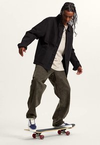 Zwarte jack met knopen over een wit shirt, gecombineerd met losse olijfgroene broek en blauwe sneakers, terwijl hij aan het skateboarden is op een houten plank.