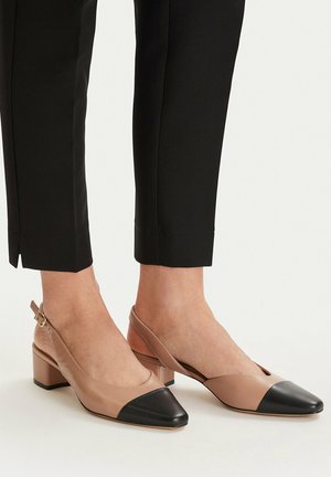 Piedi con scarpe slingback beige e nere con tacco basso abbinate a pantaloni neri corti su sfondo chiaro.