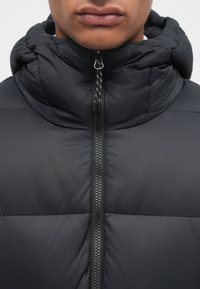 Giacca a vento nera con colletto alto, chiusura a zip e texture trapuntata. Il tessuto è liscio con una finitura leggermente lucida.