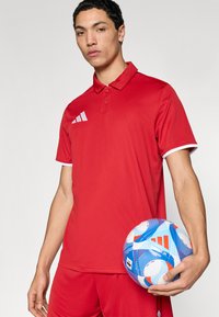 Κόκκινο πουκάμισο πόλο με λευκές λεπτομέρειες, με λογότυπο τριών ρίγων. Το μοντέλο κρατά μια μπλε και λευκή μπάλα ποδοσφαίρου με πολύχρωμα σχέδια.