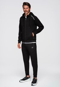 Sudadera con cremallera negra con un logo en el hombro, combinada con joggers negros que también tienen un logo. Ambos están hechos de una tela suave y tienen un ajuste relajado.