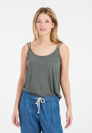 Jonge vrouw met blond haar die een grijs mouwloos topje met dubbele bandjes en blauwe broek met koord draagt, staand tegen een witte achtergrond.