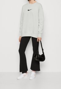 Ljust grå hoodie med svart Nike-logga, svarta ribbade utsvängda byxor, vita sneakers med rosa detaljer och en glänsande svart handväska.