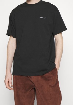 T-shirt Carhartt noir à manches courtes associé à un pantalon en velours côtelé marron, une main dans la poche, teint clair, cadré au niveau du cou et du milieu des cuisses.