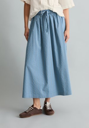 Personne portant une jupe midi à rayures verticales bleues et blanches avec un nouage à l'avant et des baskets marron à lacets blancs.