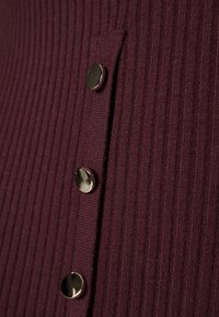 Vroubkovaná burgundská látka má řadu kovových knoflíků podél strany, což dodává detail na texturovaném povrchu.