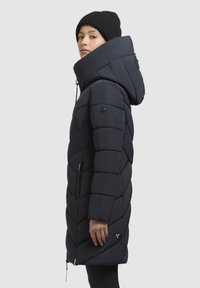 Navy pufferjack met een capuchon, voorzien van een chevron-quiltstructuur, ritszakken en een logo-accent op de zoom. Draagt een zwarte beanie.