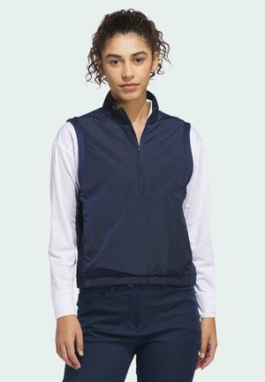 Kvinde med krøllet hår iført en navy ærmeløs vest med lynlås over en hvid langærmet skjorte og matchende navy bukser, stående mod en lys baggrund.