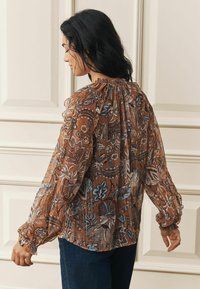 Hnedá kvetinová blúzka s dlhými, vzdušnými rukávmi a gumičkovým výstrihom. Obsahuje zložitý modrý a krémový vzor, priesvitný materiál a elastické manžety.