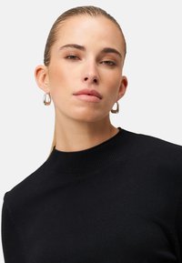 Schwarzer gestrickter Pullover mit hohem Ausschnitt. Glatte Textur, figurbetontes Design. Silberne Creolenringe ergänzen den Look.